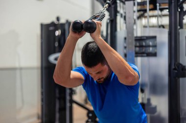 Genç fitness sporcusu, güç antrenmanı rutininin bir parçası olarak triseps egzersizleri yapmak için spor salonunda yüksek makara makinesi kullanıyor. Vücudunun üst kısımlarını geliştirmeye kendini adamış.