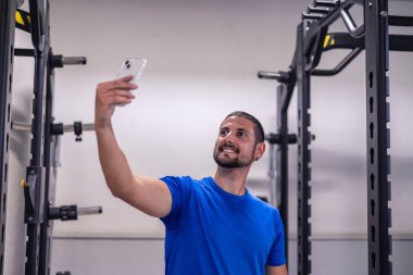 Genç sporcu spor salonunda spor yaparken sosyal medyada yayınlamak için cep telefonuyla selfie çekiyor. Fitness seyahatlerinin ve eğitim sürecinin çevrimiçi olarak paylaşılmasının modern uygulamasını vurgular