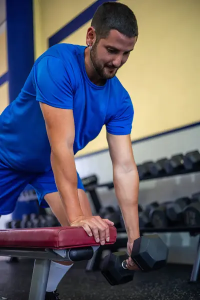 stock image vertical young athlete performs back exercises using a dumbbell while supported on a gym bench. This exercise targets the back muscles, highlighting strength training and dedication to fitness