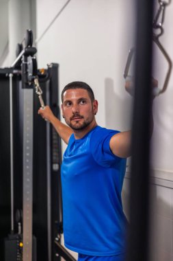 Dikey iskele adamı sırt deltoid kaslarını spor salonunda kablo makineleriyle çalıştırıyor. Bu resim sporcunun üst vücut gücünü ve kas tanımını geliştirmek için gösterdiği çabayı vurgular.