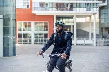 Takım elbiseli Afrikalı işadamı, şehirde elektrikli bisikletiyle dolaşırken sürdürülebilir şehir hareketliliğini vurguluyor. Görüntü, çevre dostu taşımacılıktaki kararlılığının altını çiziyor