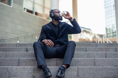 Afrikalı siyahi işadamı, şehrin finans bölgesinde merdivenlerde oturmuş kulaklıkla müzik dinleyerek kahve içiyor. Şehir yaşamındaki bir rahatlama anını tasvir eder.