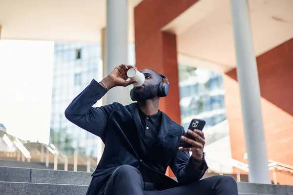 Afrika kökenli siyahi bir işadamının kahve içerken, akıllı telefonunu tutarken ve şehrin finans bölgesinde merdivenlerde oturmuş kulaklıkla müzik dinlerken düşük açılı görüntüsü.