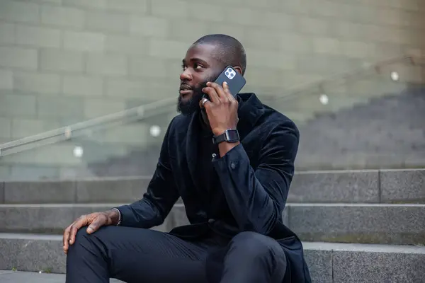 Takım elbiseli Afrikalı iş adamı, cep telefonunu kullanırken şehir merkezindeki merdivenlerde oturuyor. Fotoğraf, kentsel bir ortamda profesyonel bir ilişki anının altını çiziyor