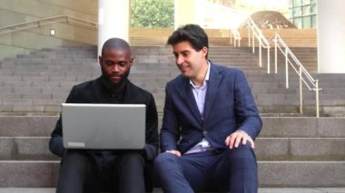İki iş adamı, biri Afrikalı Amerikalı diğeri İspanyol takım elbiseli, ofisin yakınındaki bir laptopta birbirlerine sıcak bir şekilde selam veriyorlar.