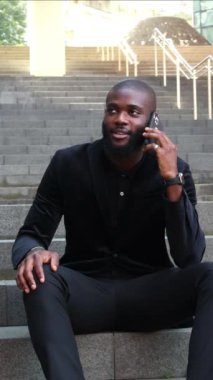 Dikey Afrikalı iş adamı ofis dışında oturuyor, iş molası sırasında cep telefonuyla konuşuyor. Sahne, profesyonel kentsel çevrede bir rahatlama ve iletişim anını yansıtıyor.