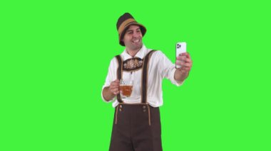 Geleneksel Oktoberfest kıyafeti giymiş Bavyeralı adam biranın tadını çıkarırken cep telefonuyla selfie çekiyor. Gülümsüyor ve Ekim Festivali 'ndeki şenlik anını yakalıyor gibi görünüyor.