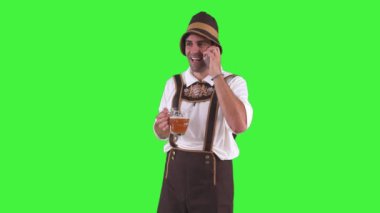 Geleneksel Oktoberfest kıyafeti giymiş Bavyeralı adam cep telefonuyla canlı bir konuşma yapıyor. Büyük bir bira bardağı krom anahtarı tutarken hareketli bir şekilde gülümsüyor.