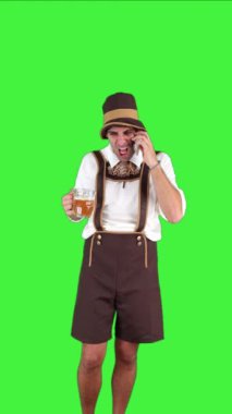 Geleneksel Oktoberfest kıyafetli dikey sinirli genç Bavyeralı elinde bira bardağıyla akıllı telefonuyla konuşuyor. Kavgacı vücut dili şenlikli arka planda göze çarpıyor.