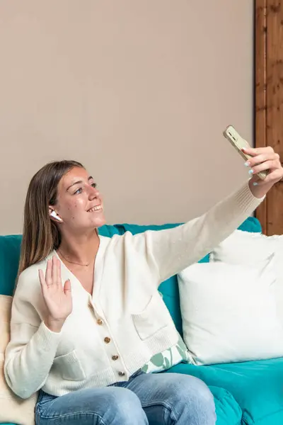 dikey sarışın kadın evdeki kanepesinde oturuyor ve akıllı telefonunu kullanarak bir video görüşmesi yapıyor. Uzaktan iletişimin rahatlığını ve modern teknolojiyi kullanmanın rahatlığını yakalıyor.