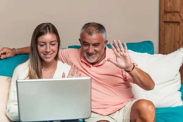 Yaşlı baba ve kızı evlerindeki kanepede oturuyorlar ve dizüstü bilgisayarlarıyla bir video görüşmesine katılırken el sallıyorlar. Görüntü sıcak ve dostane bir etkileşim yakalar.