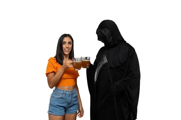 stock image grim reaper, dressed in a Halloween costume with a dark cloak and skeletal face, holds a scythe in one hand while toasting with a beer in the other alongside a cheerful young brunette woman