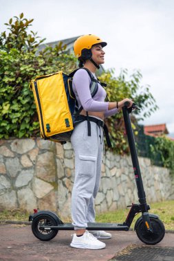 Elektrikli scooter kullanan, sarı bir sırt çantası ve miğfer takan ve emirleri ileten Latin bir kızın dikey yan görüntüsü.