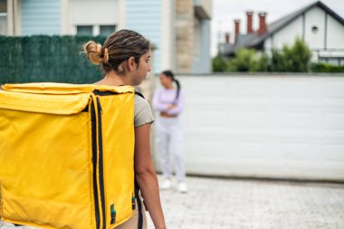 Sarı bir sırt çantası taşıyan yemek dağıtım elemanı siparişinin gecikmesi nedeniyle üzgün olan kızgın bir Latin müşterinin evine yaklaşır.