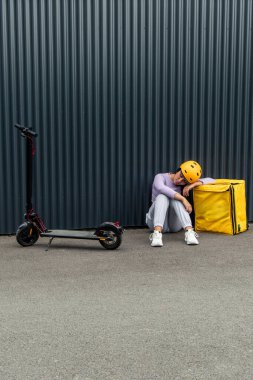 Miğfer takan dikey Latin yemek teslimatçısı kadın elektrikli scooterının yanındaki sarı sırt çantasında dinleniyor. Görüntü kısa bir soluklanma anını yakalar