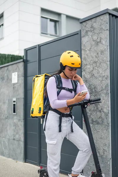 Sarı sırt çantası ve kask takan dikey Latin yemek teslimatçısı kız, elektrikli scooterıyla şehri dolaşırken yön sormak için akıllı telefonunu kullanıyor.