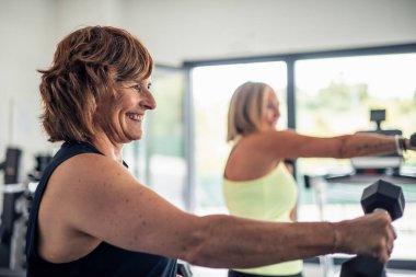 Spor salonundaki spor salonunda halterlerle kol egzersizi yaparken gülümseyen yaşlı bir kadının yakın görüntüsü. Görüntü onun zevkini ve formunu korumaya olan bağlılığını yansıtıyor.