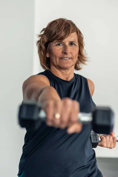 Spor kıyafetleri giymiş dikey yaşlı bir kadın spor salonundaki bir beden dövüşü sırasında kameraya bir dambılla vurmaya odaklanmış. resim onun konsantrasyonunu ve kararlılığını yakalar.