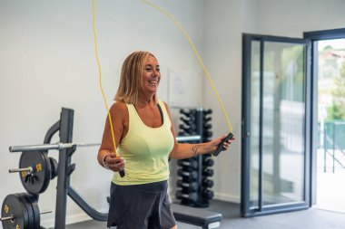Copyspace gülümseyen olgun sarışın atletik kadın spor salonundaki güç antrenmanının bir parçası olarak ip atlıyor. Atletik giysiler içinde, coşku ve enerji gösteriyor.