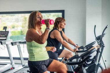 Olgun, sportif bir kadın spor salonunda dönen bir derste pembe bir su şişesinden su içiyor. Görüntü onu dinlenirken ve susuz kalırken yakalıyor. Forma olan bağlılığını vurguluyor.