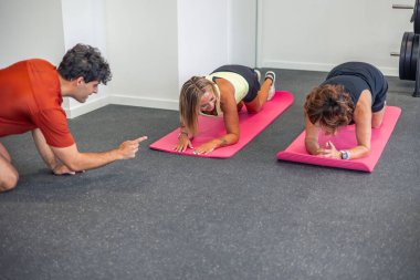 İki olgun kadın yoga minderleri üzerinde egzersiz yaparken bir koç onları spor salonunda motive ediyor. Kadınlar antrenmanlarına odaklanmış durumda ve koç onlara cesaret veriyor.