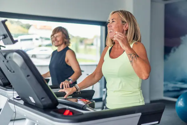Gülümseyen iki olgun spor kadın spor salonunda koşu bandında yürüyorlar, antrenmanları sırasında kardiyo çalışmaları yapıyorlar. Görüntü onların neşeli ifadelerini ve coşkularını yakalıyor.