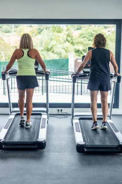 Spor salonunda koşu bandında yürürken arkadan iki yaşlı kadın görülüyor, antrenman sırasında kardiyo egzersizleri yapıyorlar. Kardiyovasküler sağlığı korumaya olan bağlılıklarını vurgular