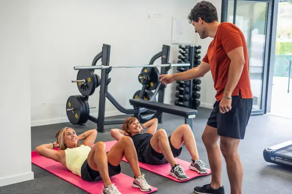 Jimnastik hocası, spor salonunda iki olgun kadını karın egzersizleri yaparken spor paspasları üzerinde cesaretlendiriyor. Kadınlar spor kıyafetlerini giydiler ve antrenmanlarına odaklandılar.