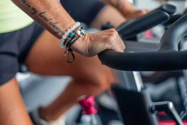 Spor salonunda bisiklet sürerken pedal çeviren yaşlı bir kadının el ve bacaklarının yakın görüntüsü. Görüntü, çalışmasındaki odaklanmasını ve çabasını vurguluyor.