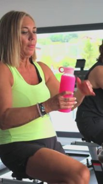 Dikey olgun atletik kadın spor salonundaki bisiklet üzerinde yaptığı egzersiz sırasında pembe bir şişeden su içerek su içiyor. Egzersizine odaklanmış ve dinç kalmaya çalışıyor.