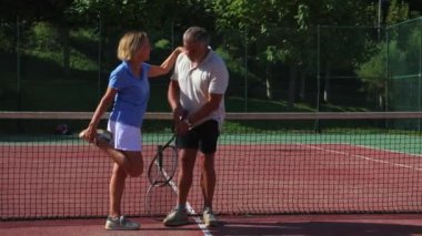 Yaşlı çift korttaki tenis antrenmanlarına başlamadan önce kas germe hareketleri yapıyor. İkisi de tenis kıyafetleri giyip yan yana duruyorlar ve vücutlarını maça hazırlıyorlar.