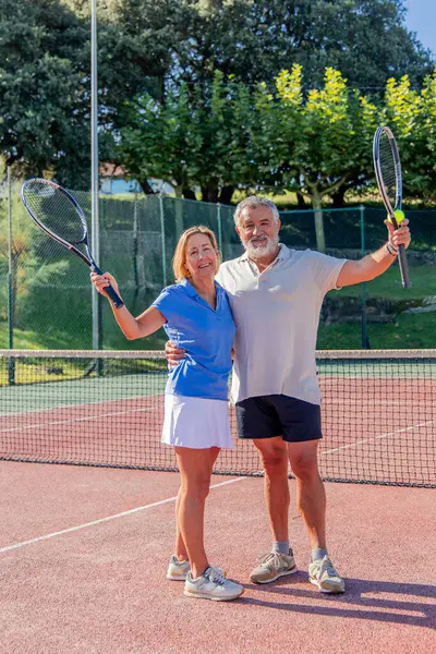 Dikey kıdemli çift sahada tenis raketlerini kaldırırken kameraya parlak bir şekilde gülümsüyor. Onların neşeli ifadeleri bir oyun sonrası başarı ve kutlama hissini yansıtır.
