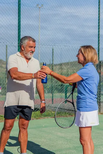 Dikey kıdemli çift güneşli bir günde tenis maçı sırasında ferahlatıcı bir su molası verir. Paylaştıkları an, spor yapmaya ve susuz kalmamaya olan bağlılıklarının altını çiziyor.