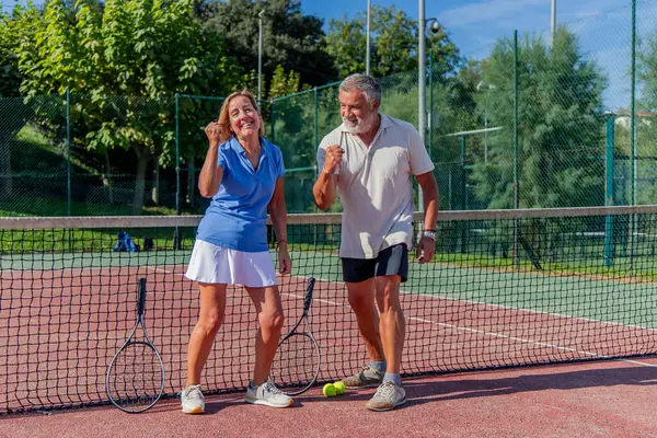 Son sınıf tenis oyuncuları maçlarını kazandıktan sonra büyük bir gülümsemeyle yumruklarını kaldırıyorlar. Onların neşeli ifadeleri başarının heyecanını yakalar.