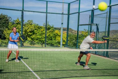 Yaşlı Padel çifti sahada aktif olarak oynuyor, topu odaklanmış ifadeler ve koordineli hareketlerle kameraya doğru gönderiyor.