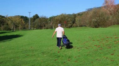 Golf arabasıyla yeşilden geçerek bir sonraki deliğe doğru giderken görülen kıdemli golfçü. Huzurlu kurs manzarası onun dikkatini ve oyuna olan bağlılığını vurguluyor.