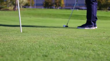 Üst düzey bir golfçüyü yakından çekerken golf sopasını dikkatle kullanıp topu yeşildeki deliğe sokuyor.