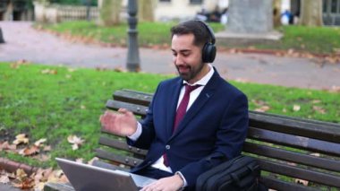 Finans bölgesinde sonbahar yapraklarıyla çevrili bir çantayla parktaki bankta otururken dizüstü bilgisayarındaki video görüşmesi sırasında resmi kıyafet dalgaları içinde iş adamı.