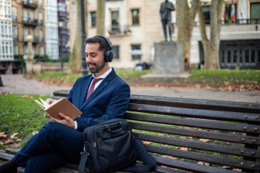 Takım elbiseli ve kravatlı mutlu bir işadamı şehir parkında bir bankta oturmuş, kulaklıkla müzik dinliyor ve kitap okuyor, mola zamanının keyfini çıkarıyor.