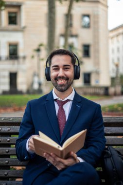 Takım elbiseli ve kravatlı genç işadamı bankta oturuyor, kulaklıklarla müzik dinliyor ve kitap okuyor, gülümsüyor ve molasının tadını çıkarıyor.