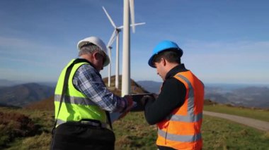 Güvenlik yeleklerindeki iki mühendis, yenilenebilir enerji gelişiminde işbirliği sergileyen açık mavi gökyüzü altında rüzgar türbinlerinin yakınındaki bir tableti incelerken proje detaylarını tartışıyor.