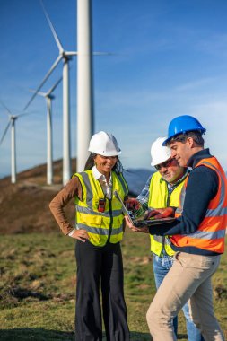 Güvenlik yelekleri ve kask giyen üç çoklu etnik mühendis bir dizüstü bilgisayar kullanıyor, rüzgar türbini santralindeki bir projede işbirliği yapıyorlar, yeşil enerjiyi kullanmak için birlikte çalışıyorlar.