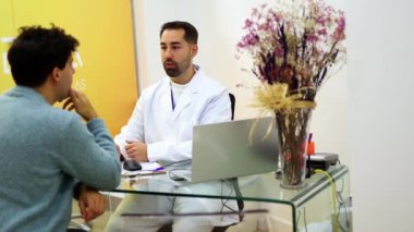 Bir masada oturan dişçi, erkek bir hastayla sohbet ediyor, elleriyle hareket ederken tedavi detaylarını anlatıyor ve rahat bir diyalog kuruyor.