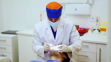 Maske takan ve eldiven takan dişçi bir hastanın dişlerini diş aletleriyle muayene eder. Hasta kapsamlı muayene sırasında rahatça bir dişçi koltuğuna uzanır.