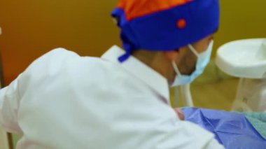 Maske ve cerrahi başlık takan dişçi, dişçi koltuğunda uzanan ve modern ekipman ve parlak klinik aydınlatmalarıyla çevrili bir hastaya diş prosedürü uyguluyor.