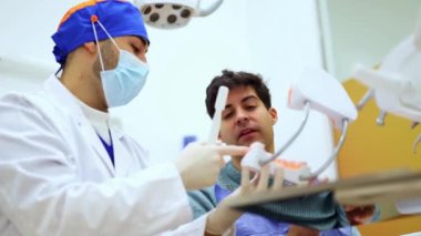Maske takan ve cerrahi başlık takan dişçi erkek hastaya diş fırçalamanın doğru yolunu açıklıyor. Diş kalıbı kullanıyor.