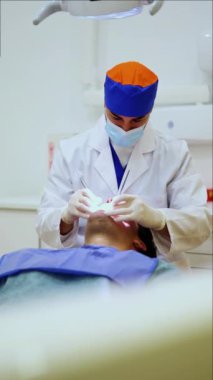 Maske ve eldiven takan dikey dişçi bir diş kliniğinde hastanın dişlerini muayene eder. Hasta, temiz, steril ekipman ve parlak ışıklandırma ile çevrili bir diş sandalyesine yaslanıyor.
