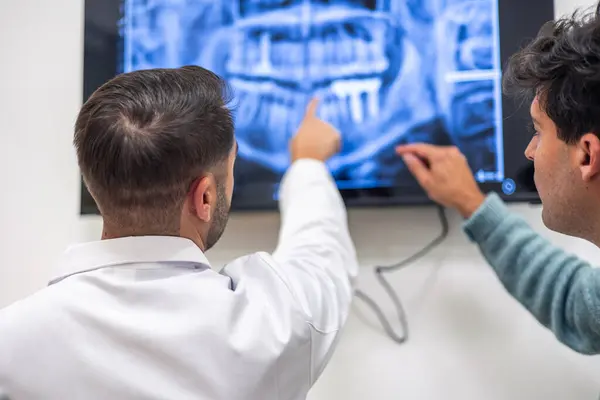 Profesyonel dişçi hastasına panoramik diş röntgenini göstererek modern bir diş kliniğinde teşhis ve tedavi planını açıklıyor.