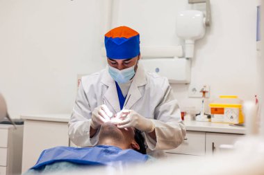 Cerrahi maske ve eldiven takan dişçi modern klinikte dişçi koltuğunda uzanan erkek hastaya diş aletleri ve ekipman kullanarak diş kontrolü yapıyor.