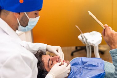 Maske takan ve eldiven takan dişçi modern diş kliniğinde diş aletlerini tutarken hastanın dişlerini muayene ediyor.